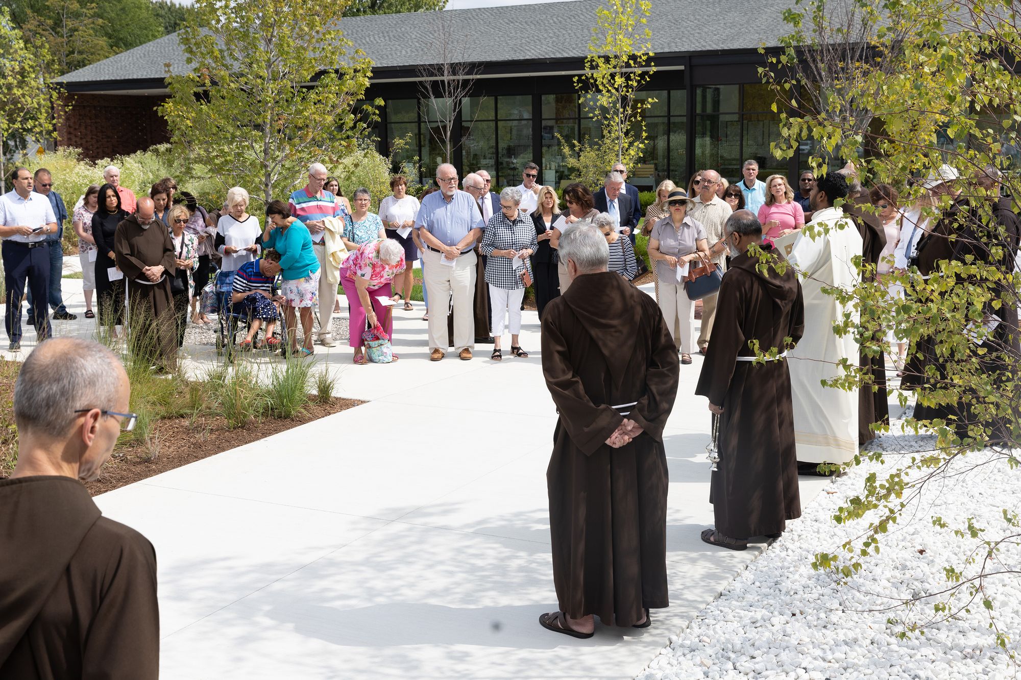 A landscape color photograph of a crowd of friars, benefactors and other supporters pray during the blessing and dedication of the Solanus Casey Center's new rosary garden, votive chapel and outdoor space.