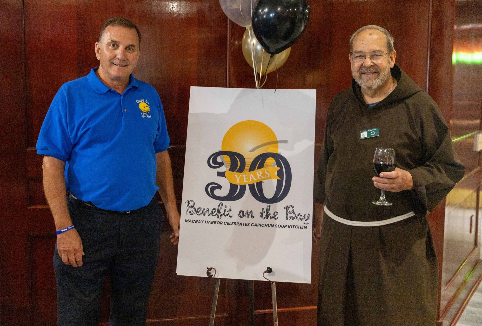 A color landscape photograph with a sign on an easel at center that reads "30 Years of Benefit on the Bay. Macray Harbor celebrates Capuchin Soup Kitchen." The sign is flanked by Shelving Inc. President Mike Schodowski at left and Fr. David Preuss, OFM Cap. at right.