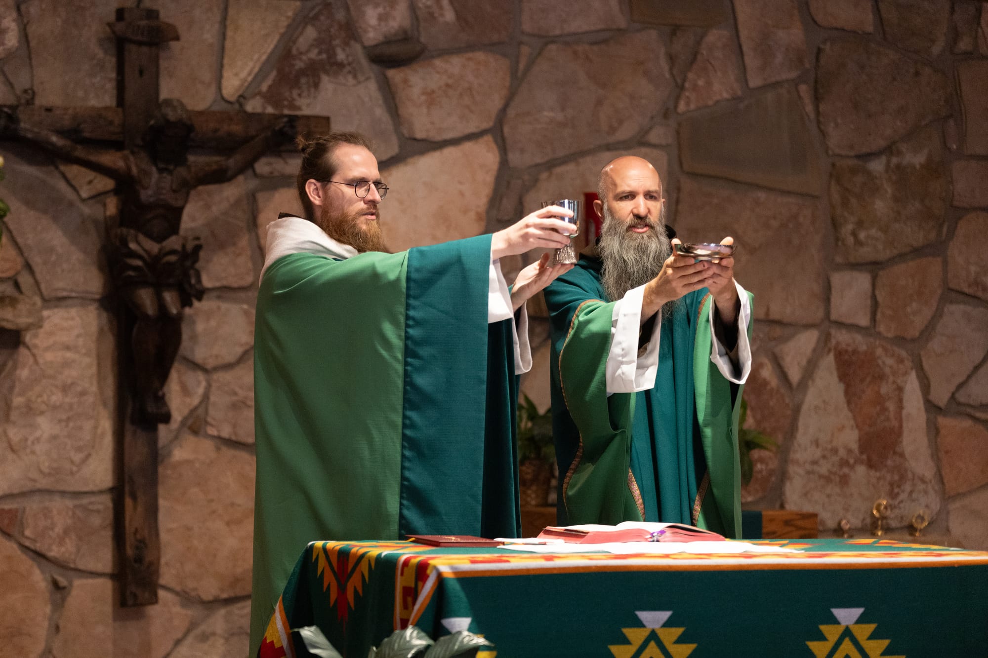 Friars Thomas Skowron and Mike Dorn elevate the body and blood of Jesus during Mass at the Crow Parishes. They are wearing green vestments signifying that it is Ordinary Time.
