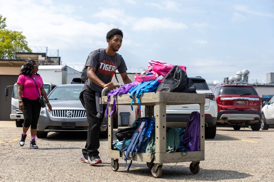 A color landscape photo of a volunteer moving a cart loaded with backpacks during the back-to-school drive distribution.